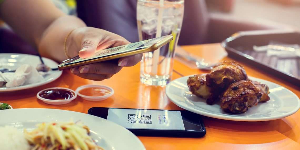 Selective focus to smartphone in hand to scan QR code on tag with blurry food, dessert and customers in restaurant to accepted generate digital pay without money. Qr code payment concept.