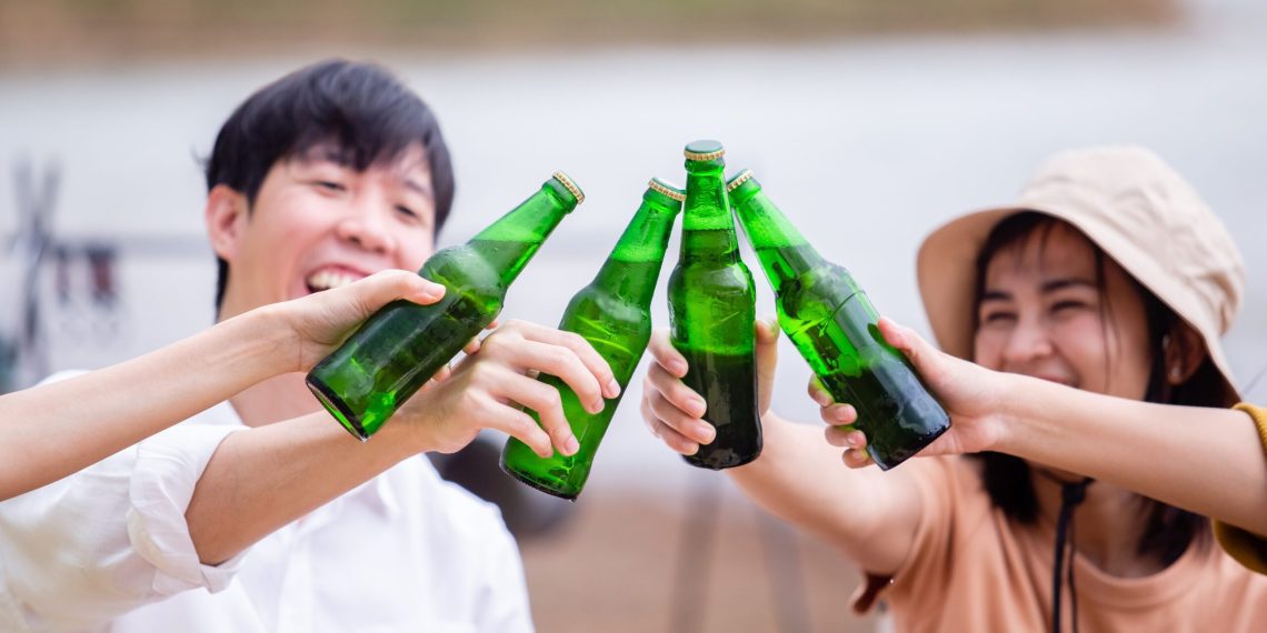 Group of young Asian friends party on vacation enjoy drinking at camping lake side. close up cheering with beer bottle. hands holding alcoholic beverages. Picnic in holiday.